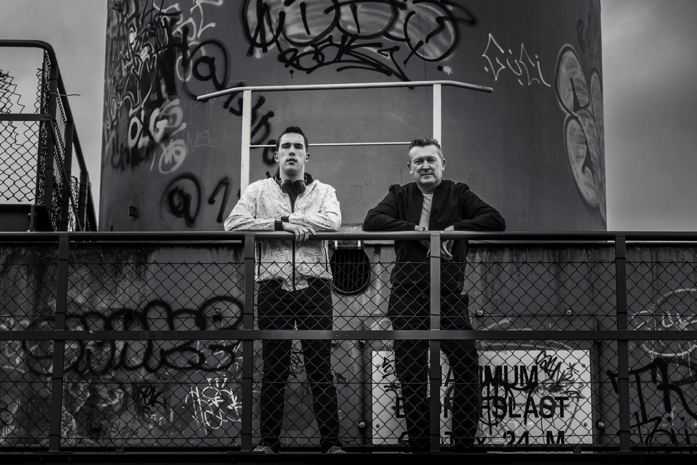 Miky and Sebasto V leaning on a metal railing in front of a graffiti-covered concrete tower, in black and white