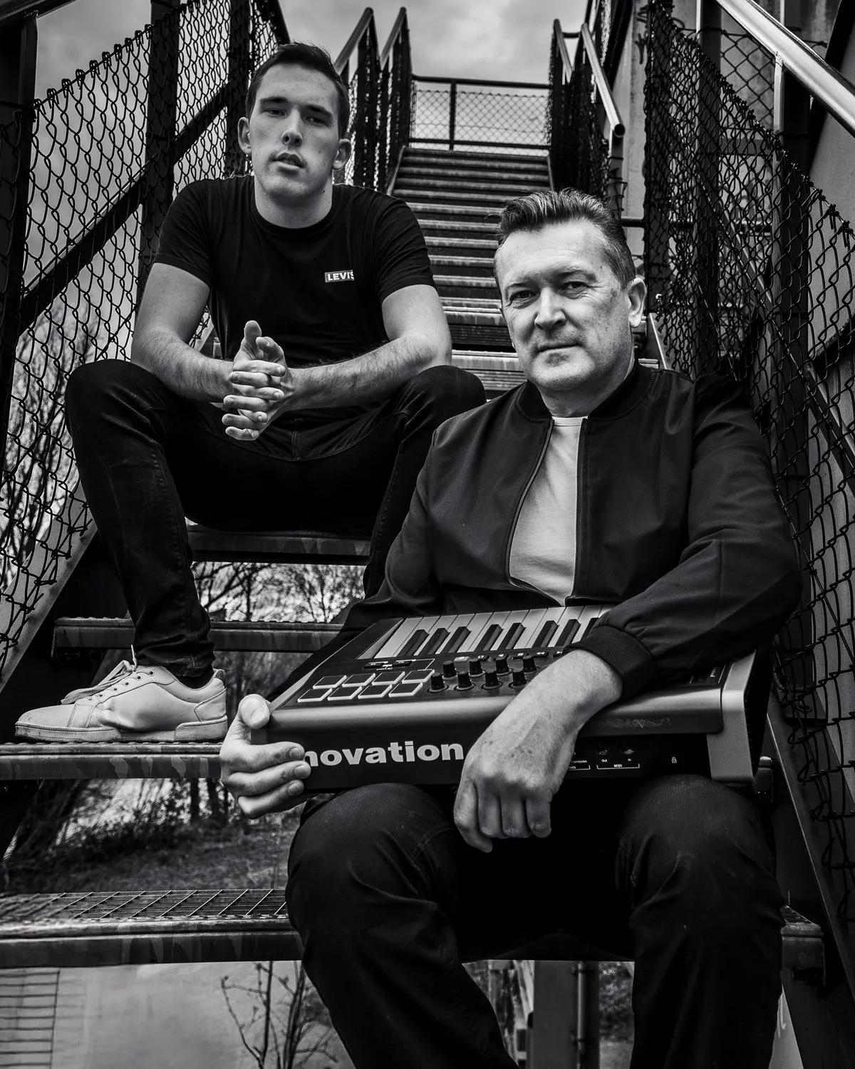 Miky and Sebasto V sitting on an outdoor steel staircase, one of them holding a Novation keyboard, in black and white